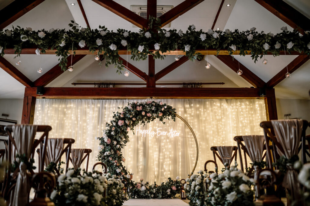 Image of an indoor wedding ceremony set up with a backdrop star cloth with hundreds of sparkling white lights and a neon sign in a circular floral backdrop