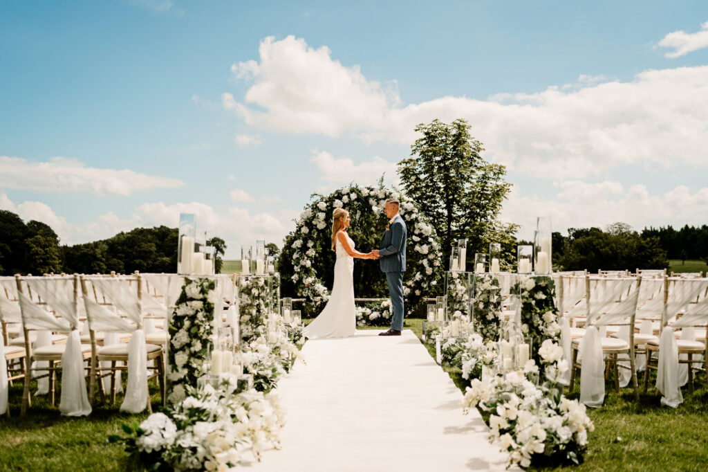 Outside wedding ceremony, with a white carpet aisle, chairs either side of the aisle and a large circular floral backdrop with the bride and groom standing staring lovingly into each others eyes.