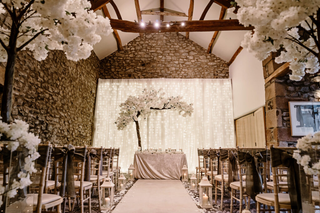 Winston Barn at The Coniston set up for a wedding ceremony with aisle and chairs either side and a large tree with white leaves behind the registrars table